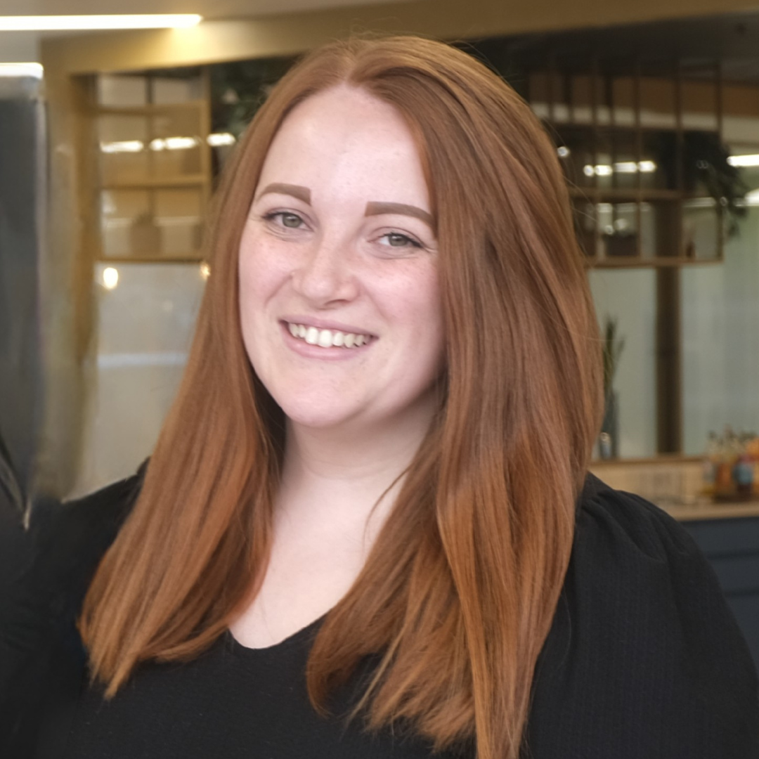 Smiling woman with long red hair in a black top, standing indoors with a blurred office background