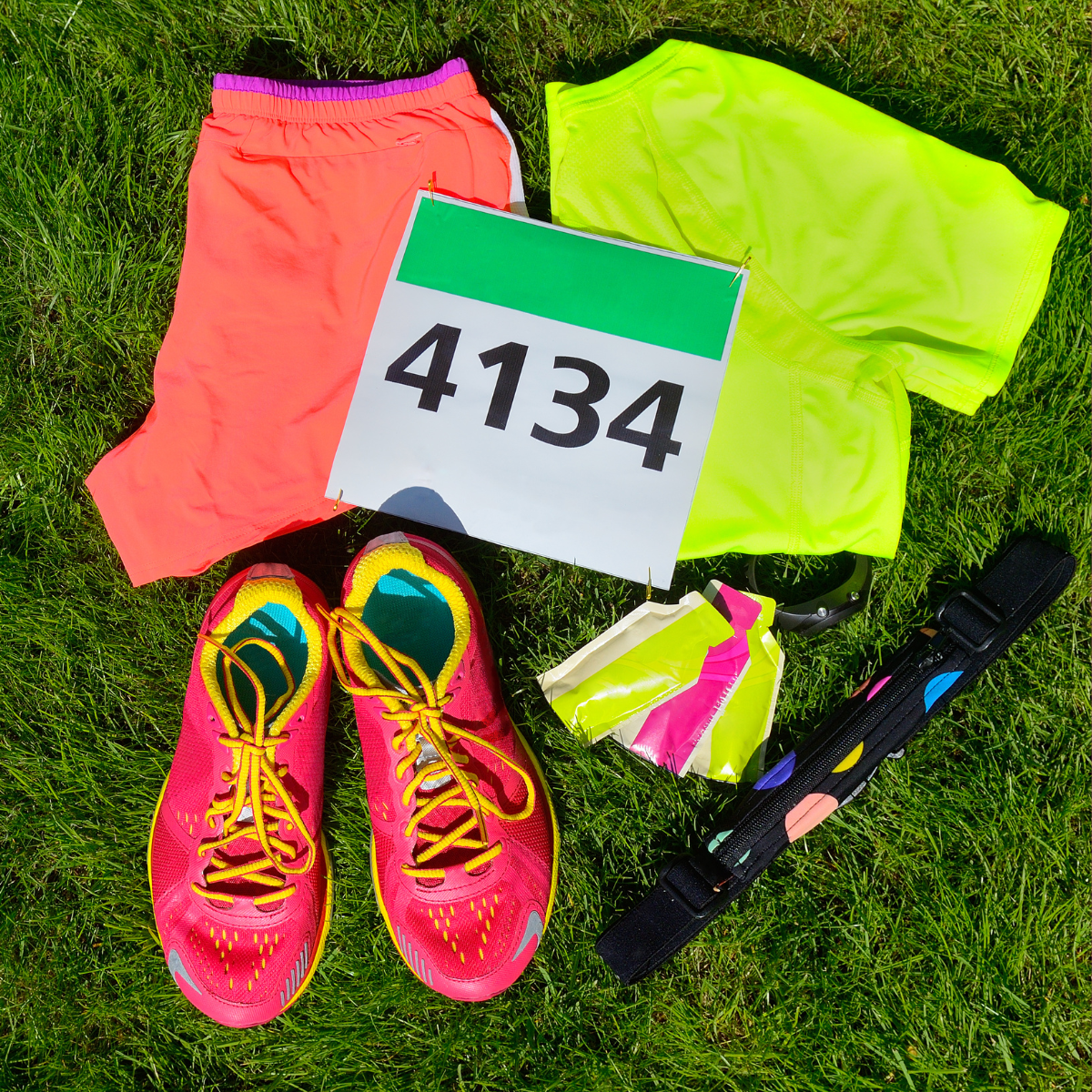 Running gear laid out on grass: neon shorts, race bib 4134, pink shoes, and a black baton.