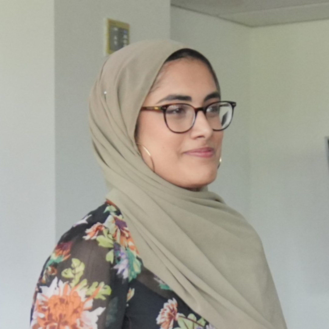 Person in glasses and beige hijab wearing a floral top, standing indoors against a light wall