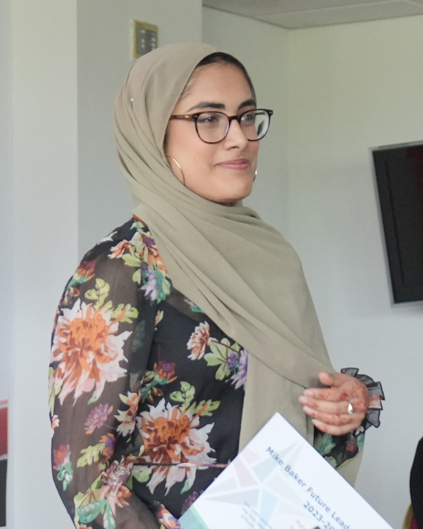 Woman in beige hijab and glasses holding papers, wearing a floral dress indoors.
