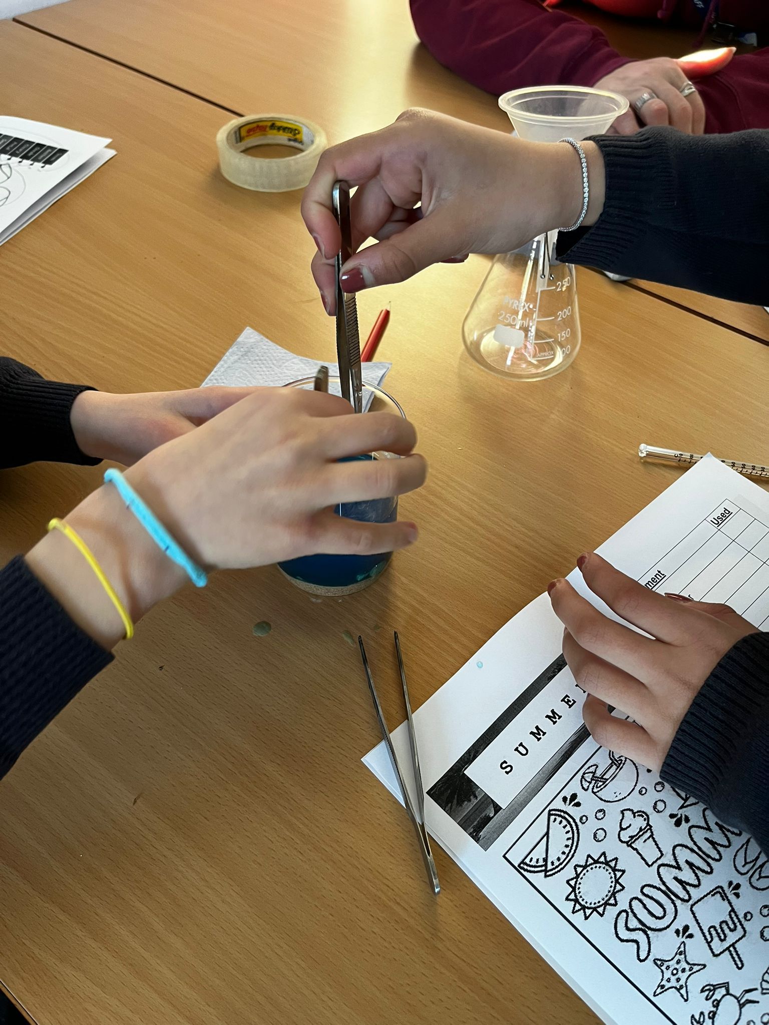 Hands around a table use tools to measure liquid into a beaker during a classroom science activity