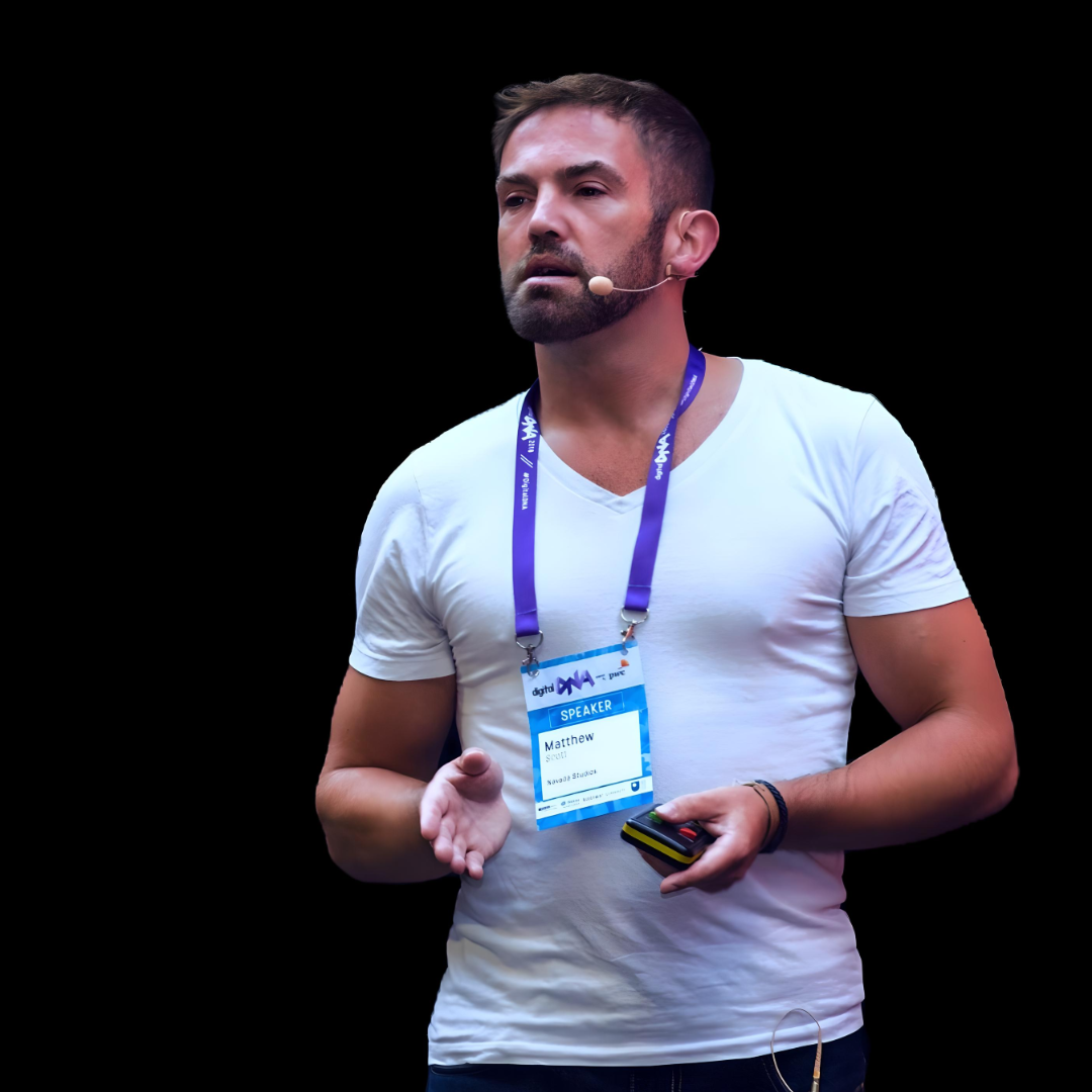 Man speaking on stage in a white T-shirt, wearing a lanyard and holding a phone
