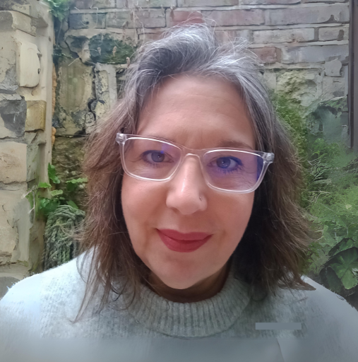 Portrait of a person wearing glasses and a light sweater, standing before a brick wall and greenery.