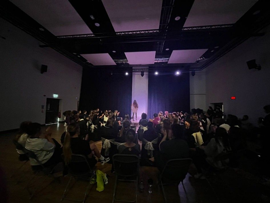 Audience seated in a dim hall facing a stage with purple lights and a white backdrop