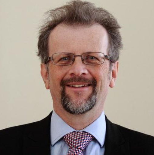 Portrait of a smiling man in glasses, suit, and patterned tie against a plain background