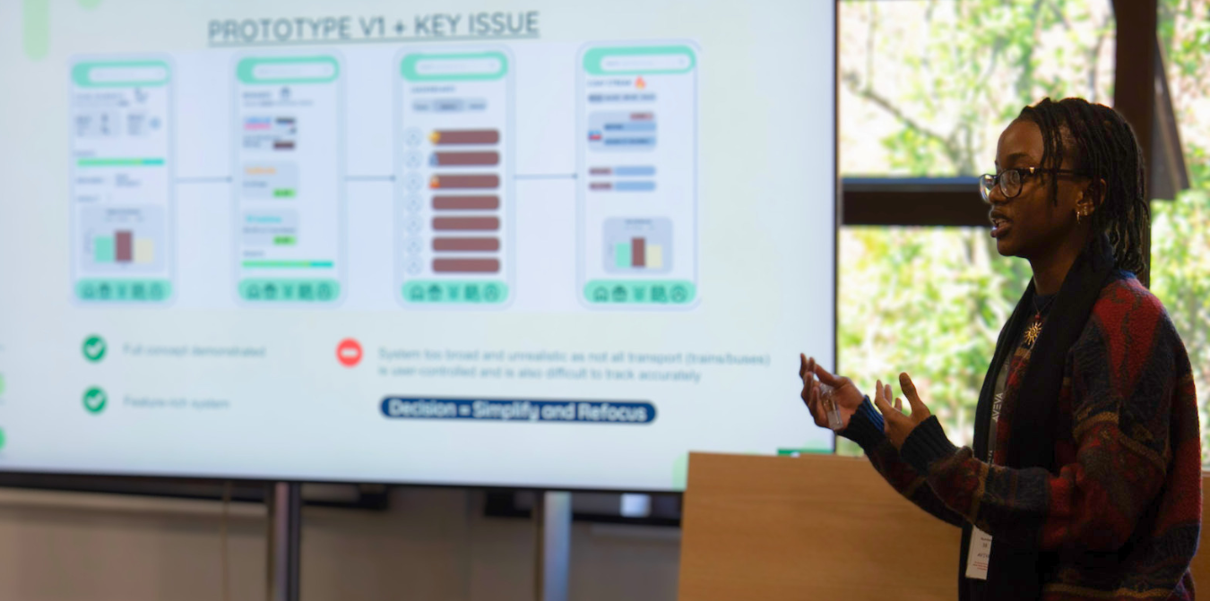 Young student presenter speaking beside projected slides in a meeting room, with colourful diagrams on the screen