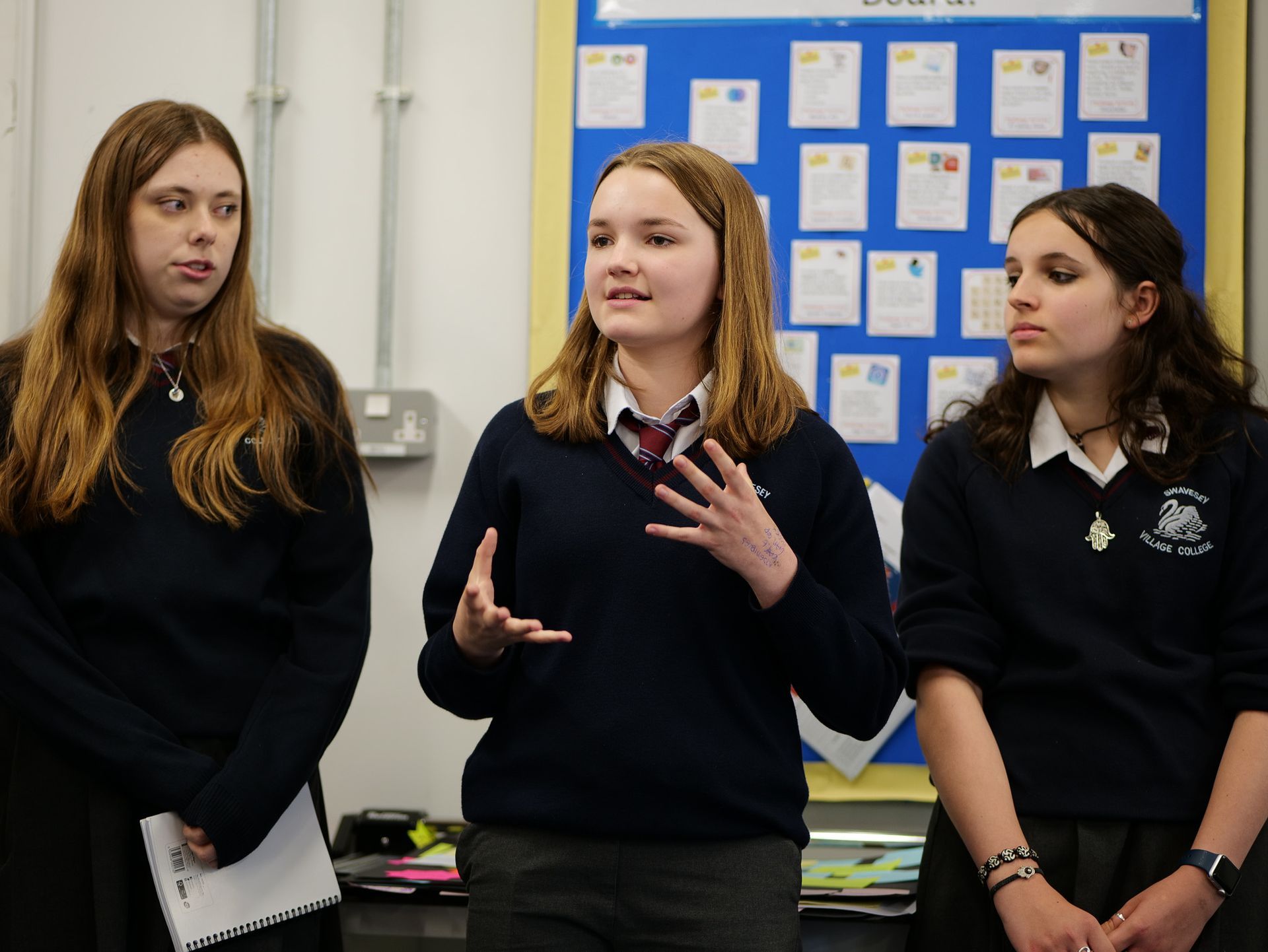 Three students in dark uniforms stand speaking in a classroom beside a bulletin board.