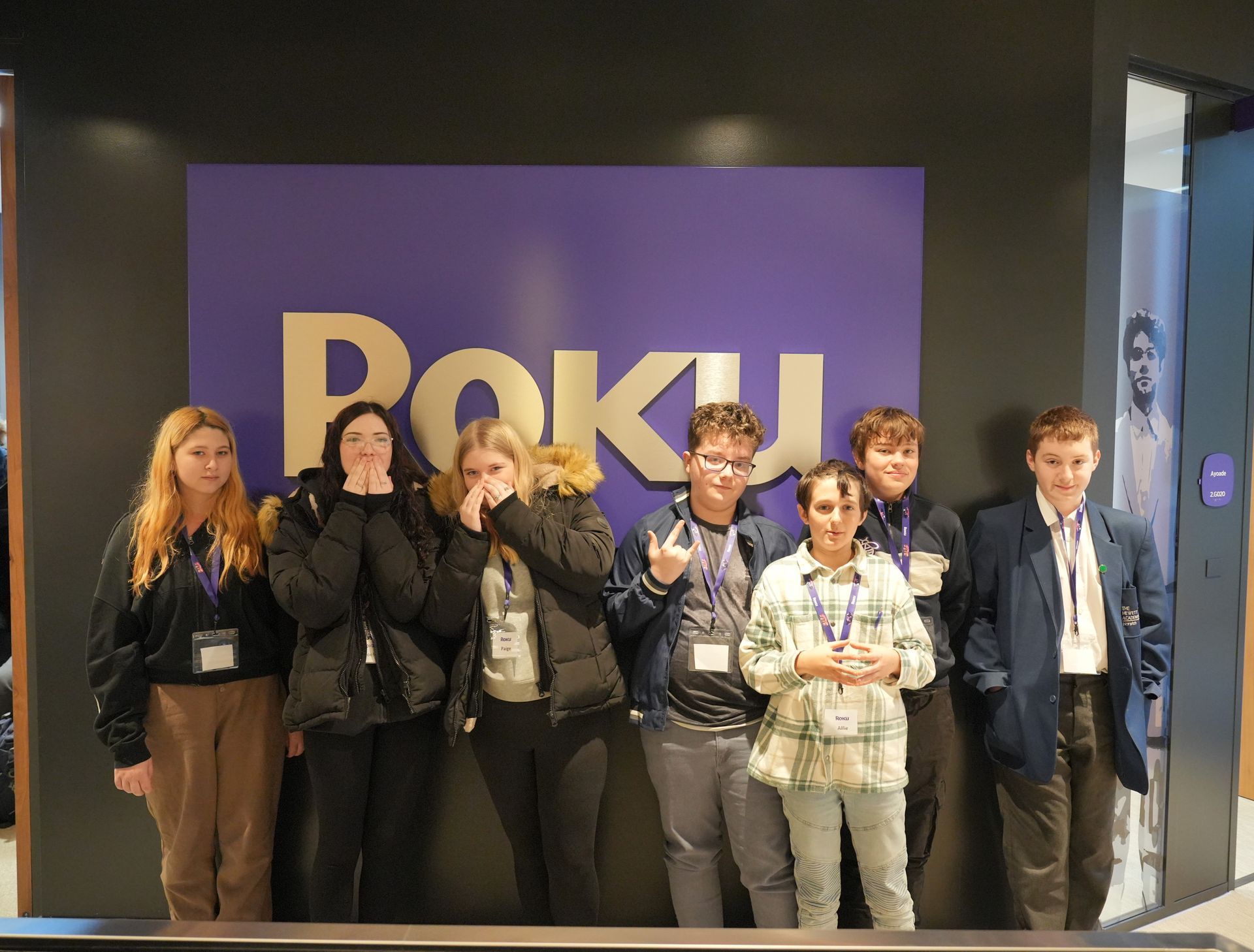 Group posing in front of a purple “ROKU” display at an event booth, smiling and waving.