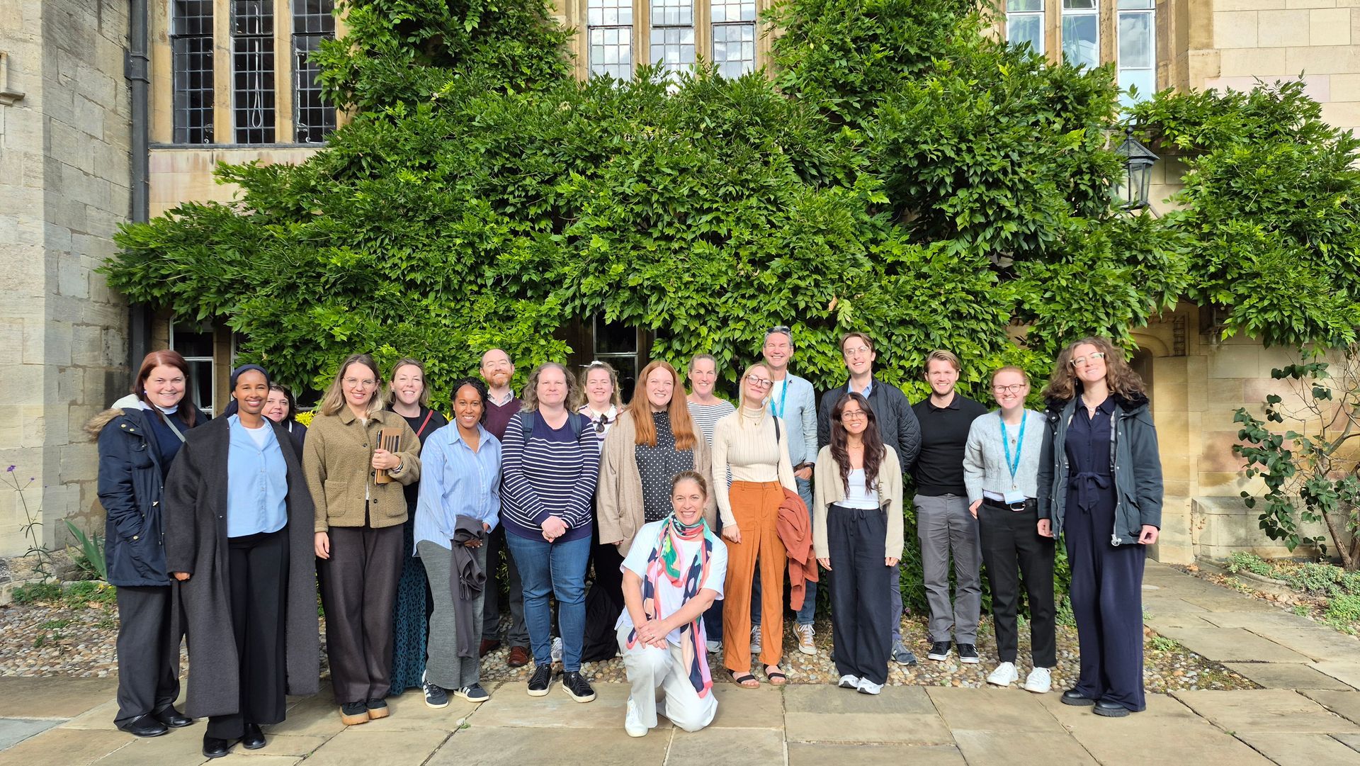 Group of people standing outside a stone building in front of a large green tree, posing for a photo.