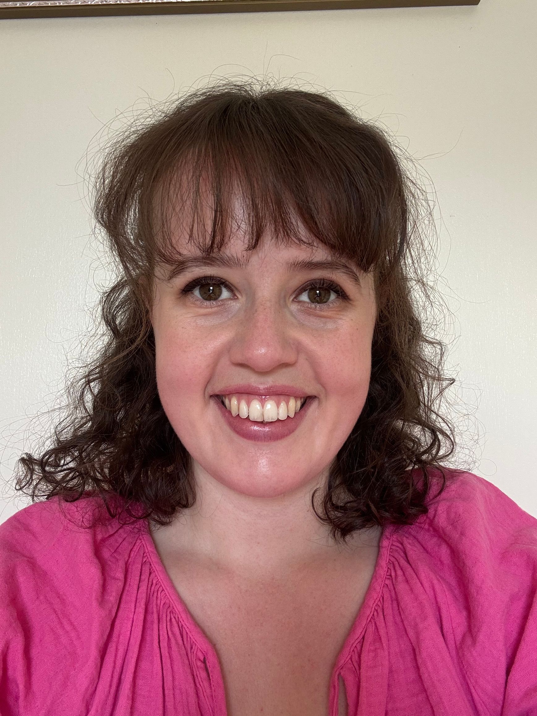 Smiling woman with curly brown hair wearing a bright pink top against a plain wall