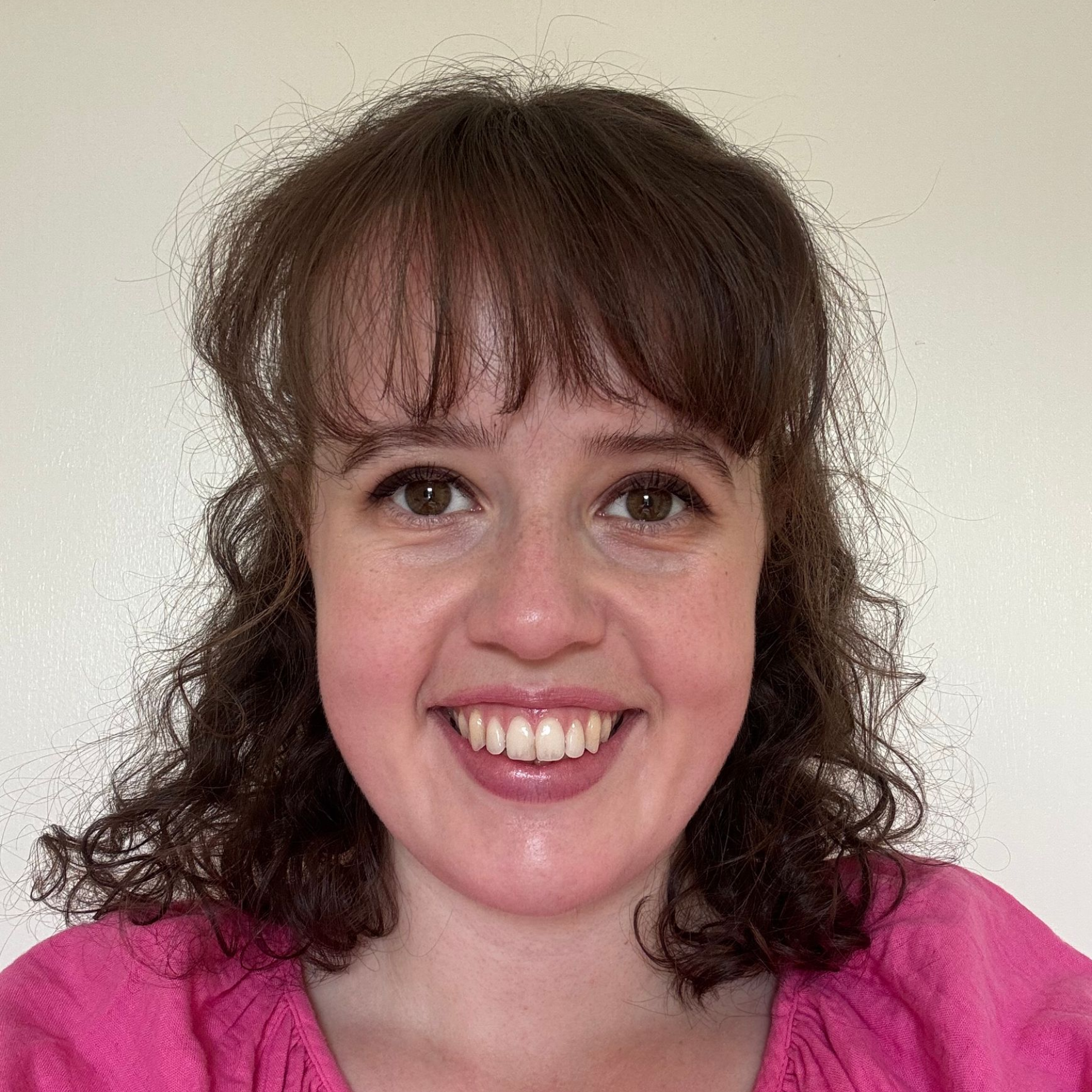 Smiling woman with curly brown hair and bangs, wearing a pink top against a plain white background