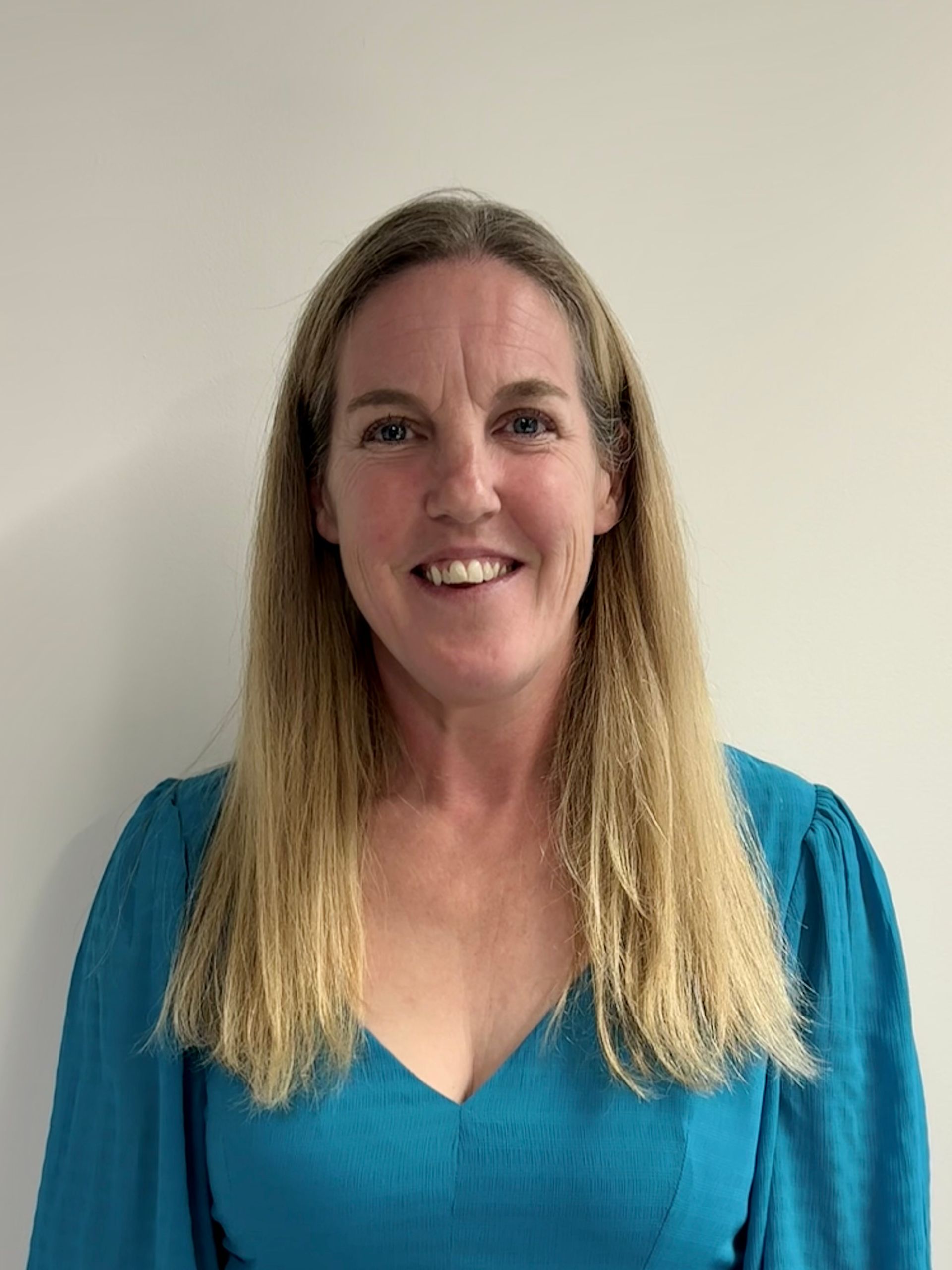 Smiling woman in a teal top standing against a plain light wall.