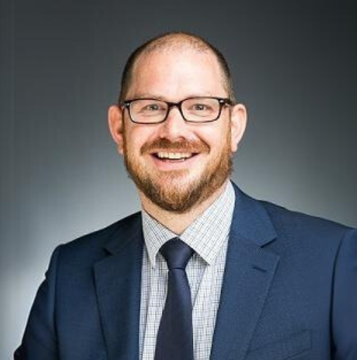 Professional headshot of a smiling man in glasses, navy suit, and tie against a gray background