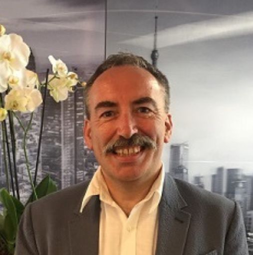 Smiling man in a gray suit standing indoors, with white orchids and city skyline wall art behind him