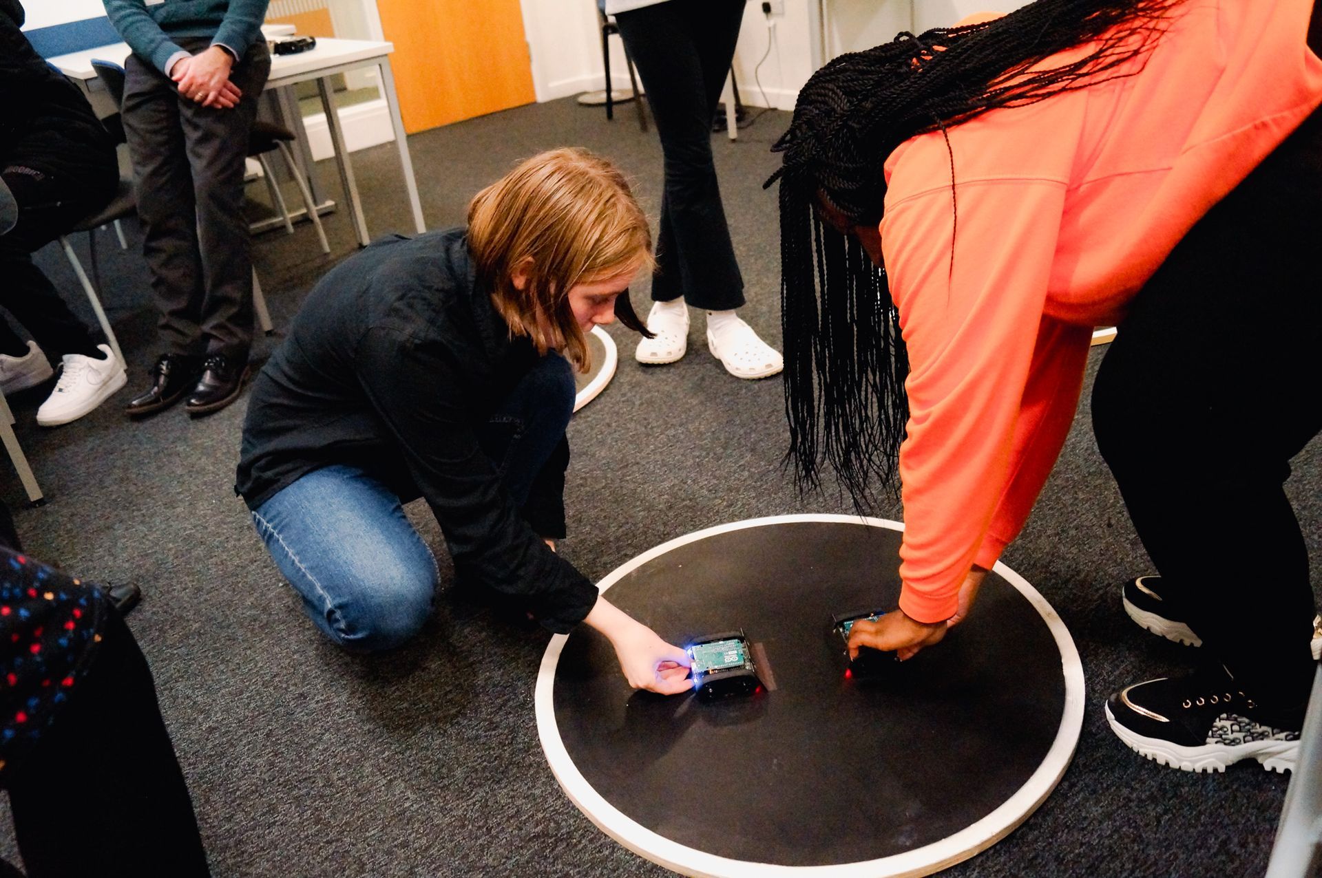 Two people crouch over a white circle on a carpet, using a smartphone and touching a small object.