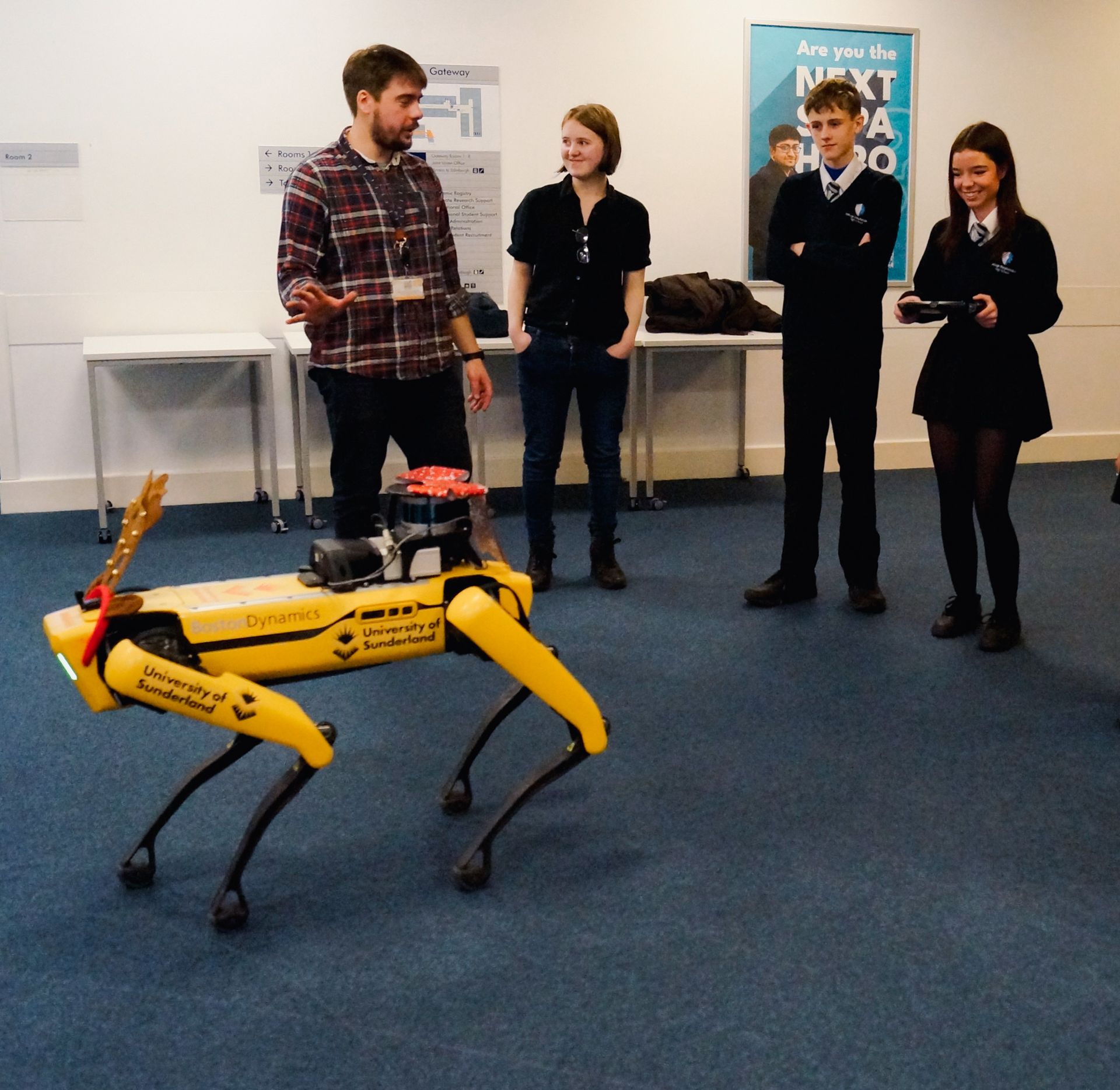 Yellow robot dog on blue carpet beside three people in a room, with posters on the wall.