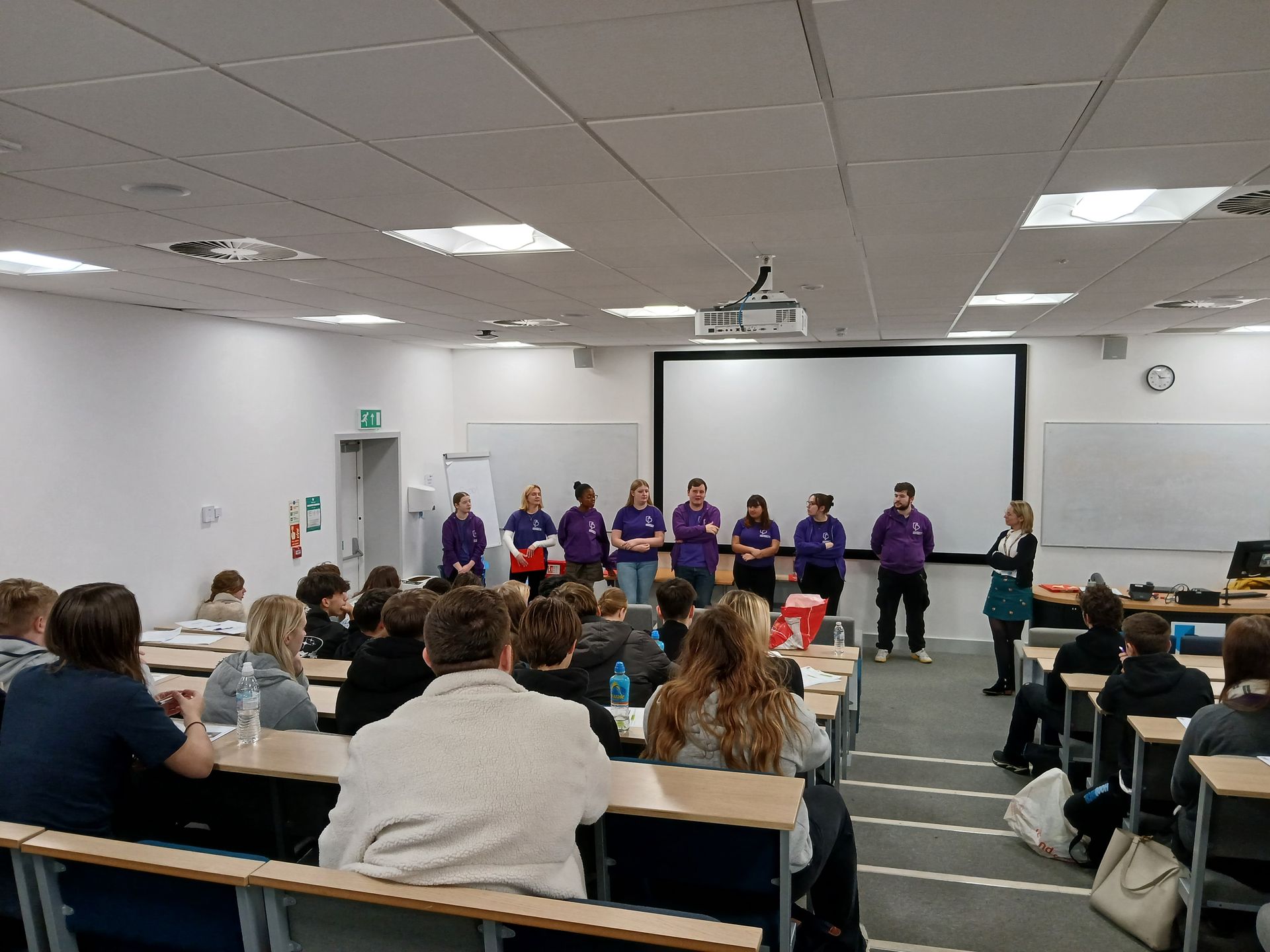 Audience watches a group in purple shirts presenting at the front of a conference room