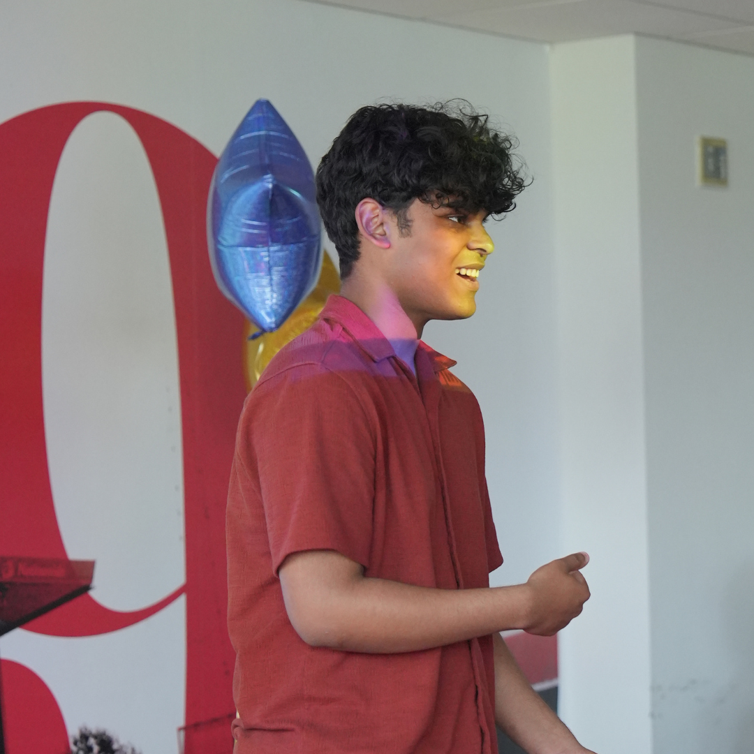 Person in a red shirt standing indoors beside a blue balloon, in front of a red and white backdrop.