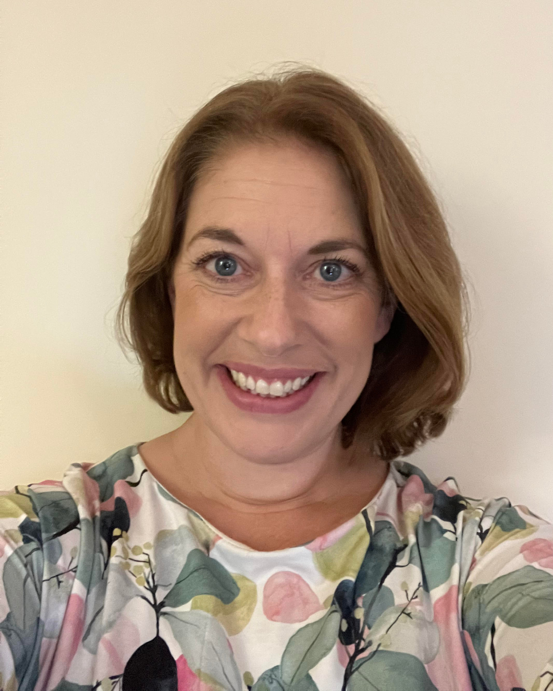 Smiling person in a floral top against a plain beige wall