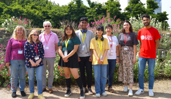 Group of people standing outdoors in a garden, smiling and posing together in bright casual clothing.