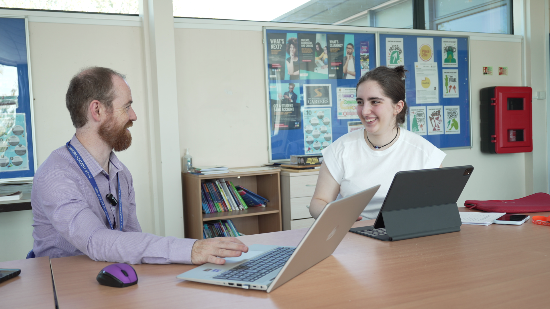 Two people meeting at a table with laptops in a classroom or office setting.