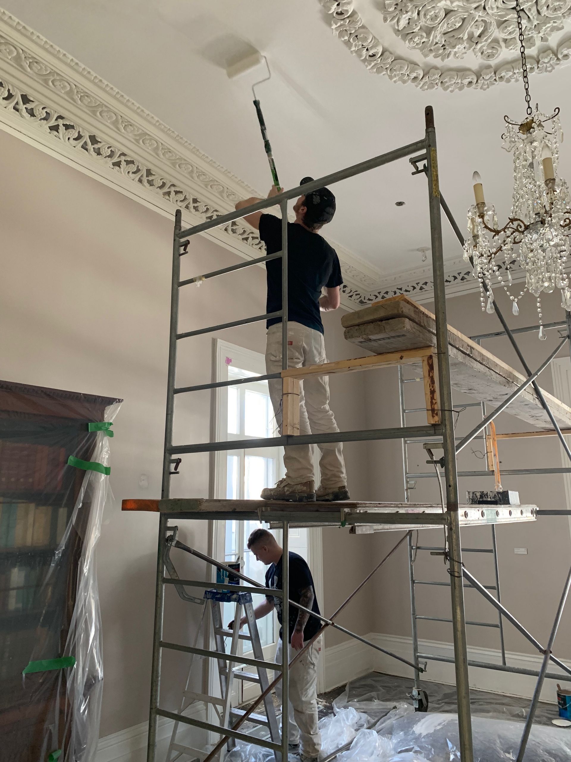 Two men are painting the ceiling of a room on a scaffolding.
