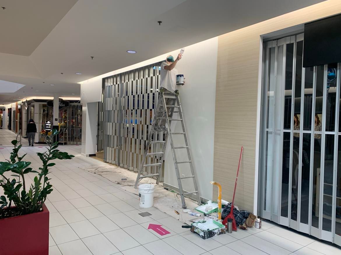 A man is standing on a ladder painting a wall in a mall.