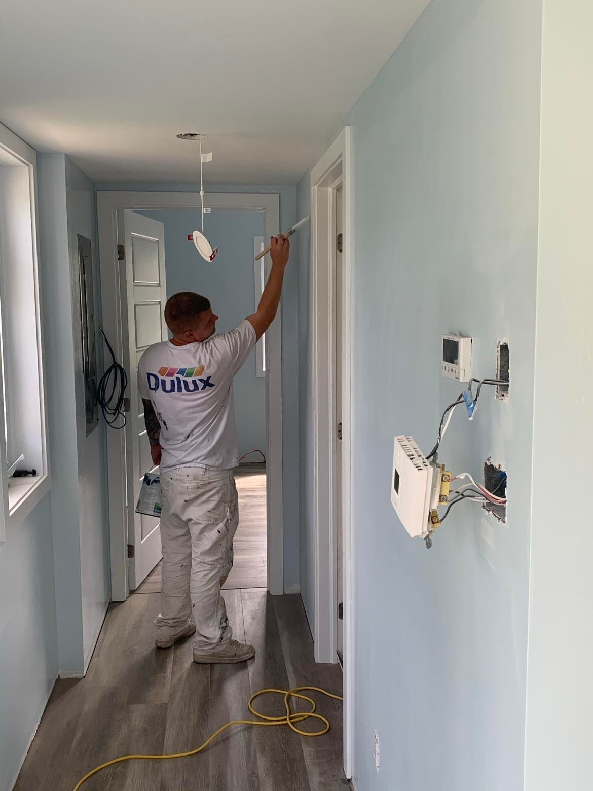 Person painting a light blue hallway with white trim; unfinished electrical work visible.