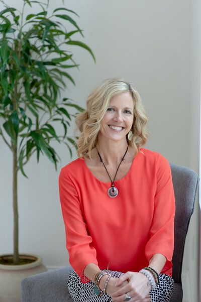 Smiling woman in a coral top seated indoors beside a potted plant