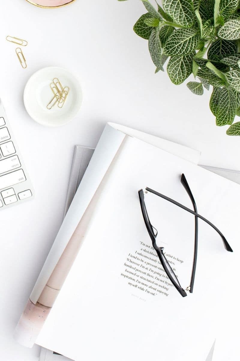 Minimal white desk with open book, eyeglasses, white candle, and green plant.