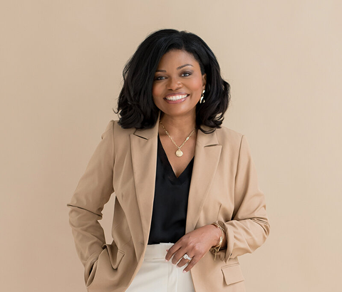 Smiling person in a tan blazer and black top, standing against a plain beige background.