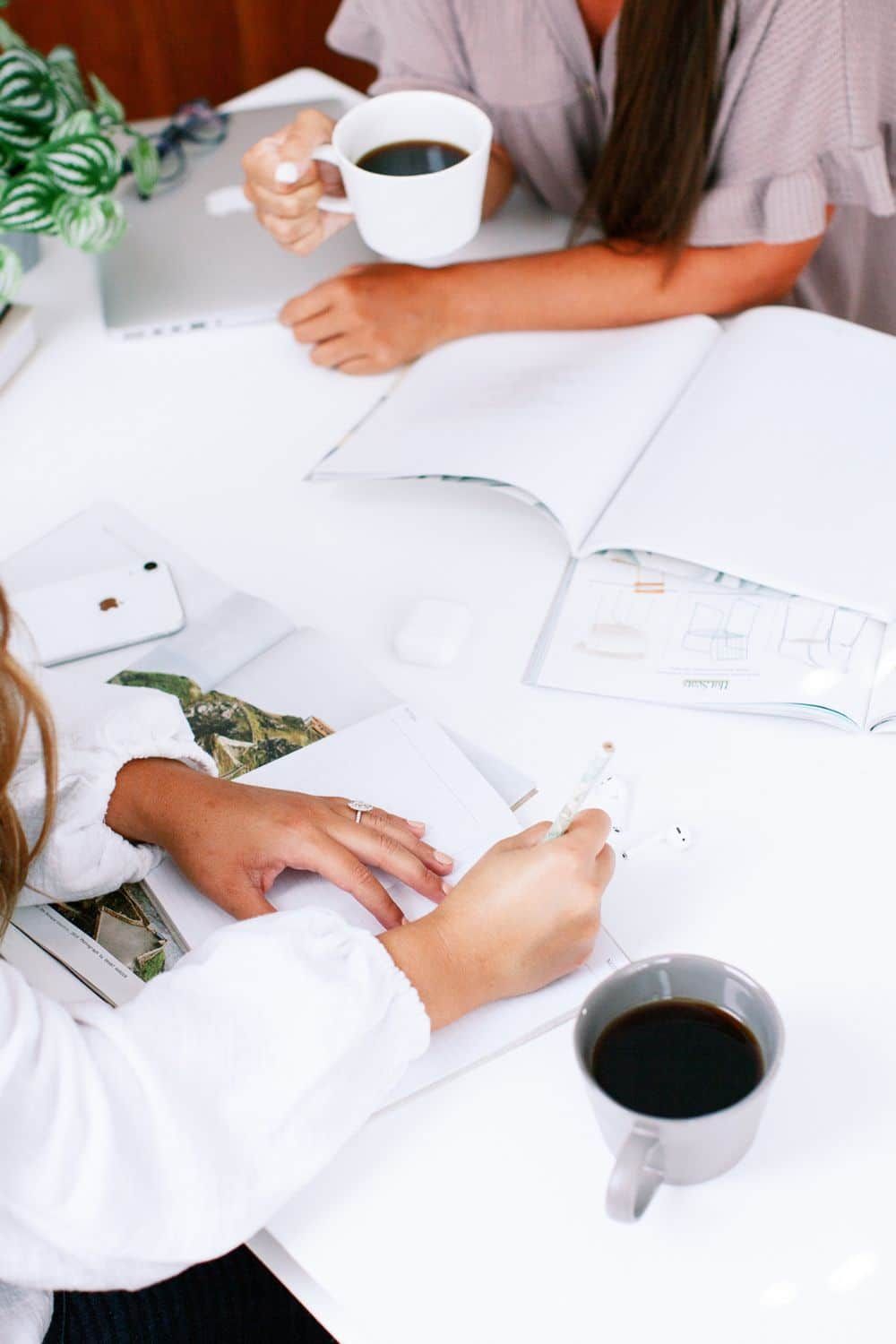 People writing and drinking coffee around a white table with open notebooks and papers
