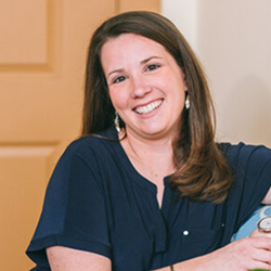 Smiling woman in a navy top, seated indoors beside a wooden door