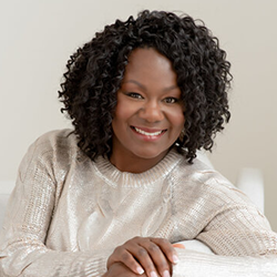 Smiling person with curly hair in a cream sweater, leaning on a white chair in a bright room