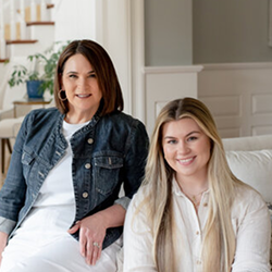 Two women smiling on a white sofa in a bright living room, one in denim jacket and one in a white blouse