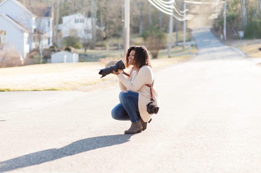 Person crouching on a sunlit street, holding a camera and taking a photo.