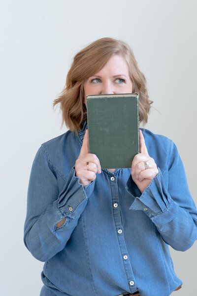 Woman in a blue shirt peeks over a small green notebook against a light background