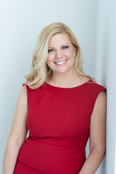 Smiling blonde woman in a red sleeveless dress against a light blue background