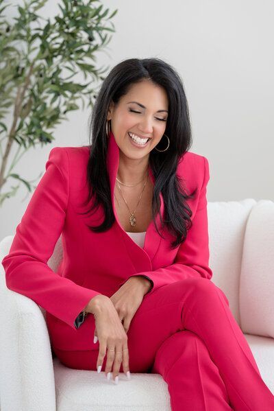 Woman in a bright pink suit smiling on a white sofa beside a potted plant