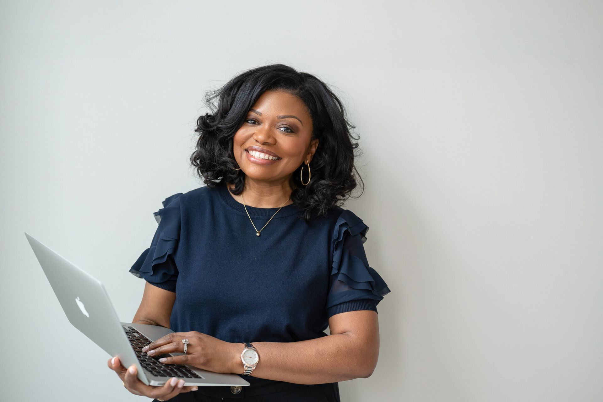 Woman in a burgundy blouse smiling while using a laptop at a bright office desk