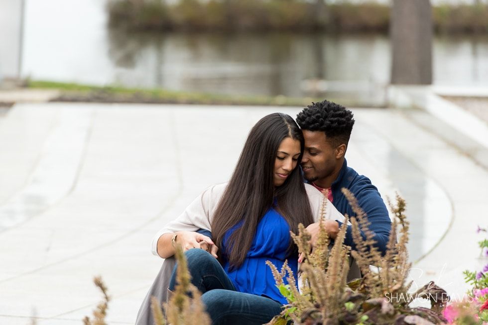 Couple sitting close together on a curved walkway by flowers, with a blurred fountain in the background