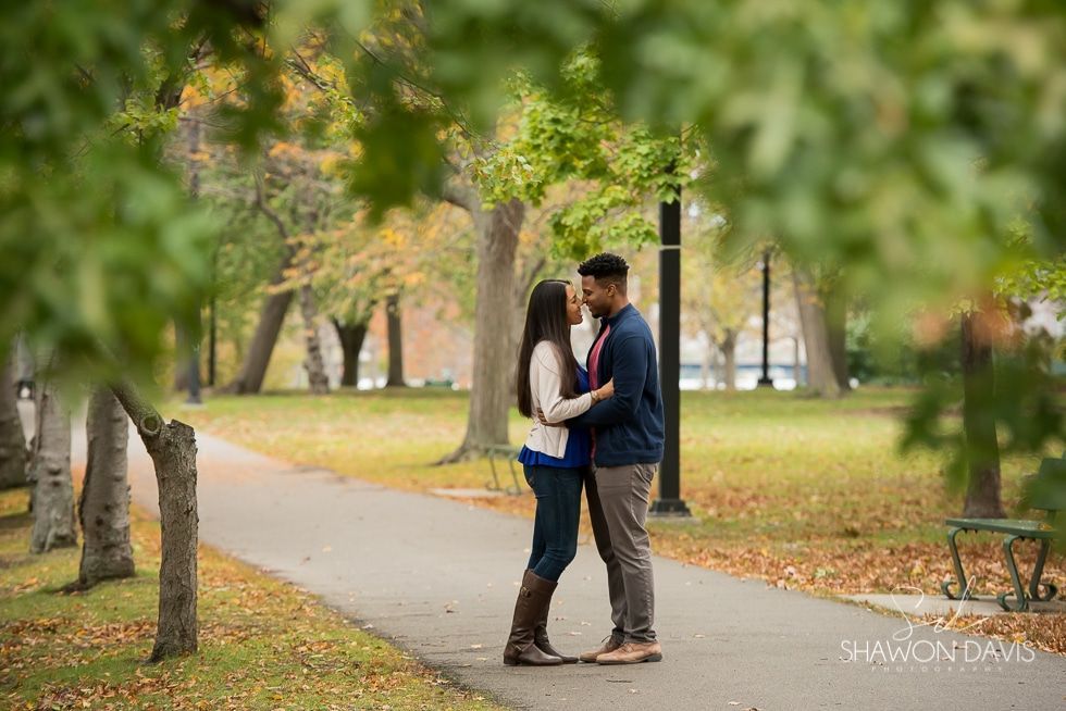 Couple embracing on a tree-lined park path in autumn, with blurred green leaves in the foreground.