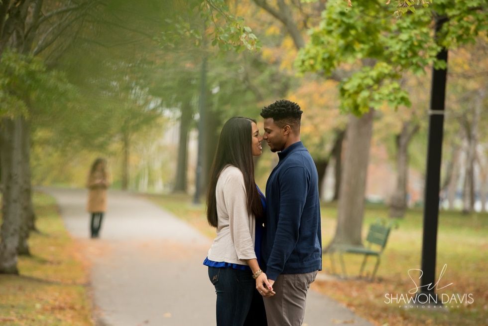 Couple standing face to face on a tree-lined path in a park, with a blurred person in the background