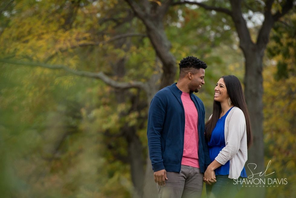 Couple smiling at each other in a park, standing among trees in blue, pink, and white clothing