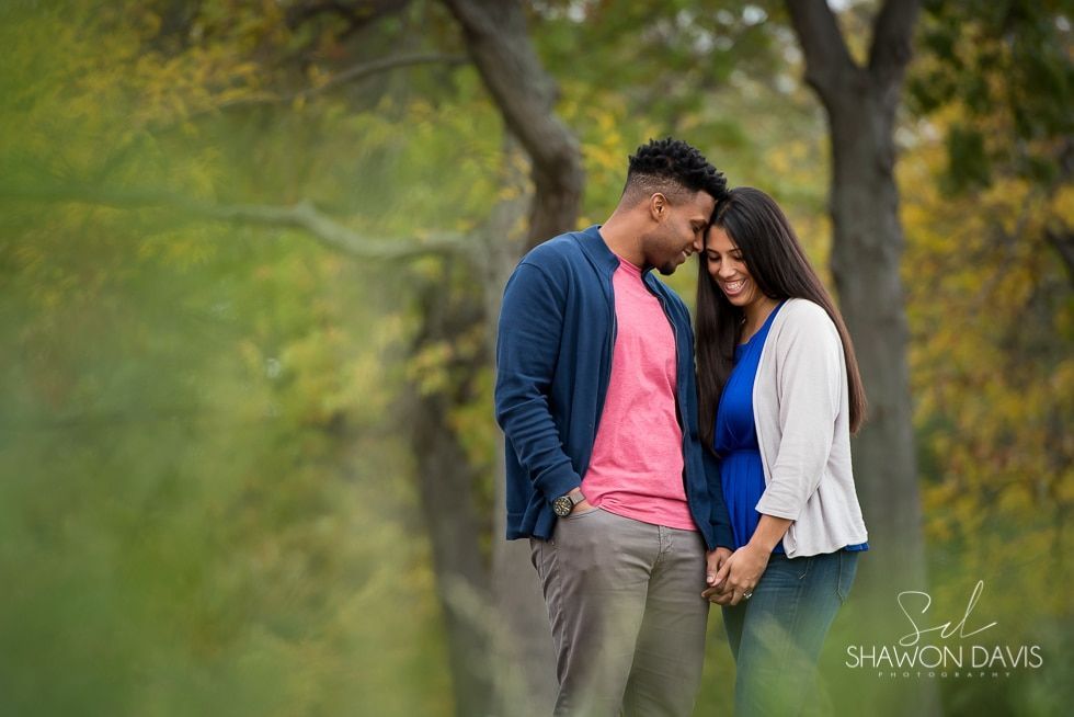 A couple stands close together in a soft-focus outdoor park setting, smiling and leaning their foreheads together.