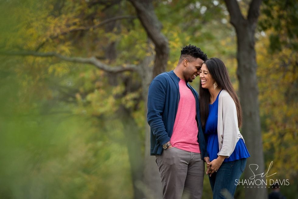 Couple standing close together in a blurred park, smiling with foreheads touching, wearing pink, blue, and white.