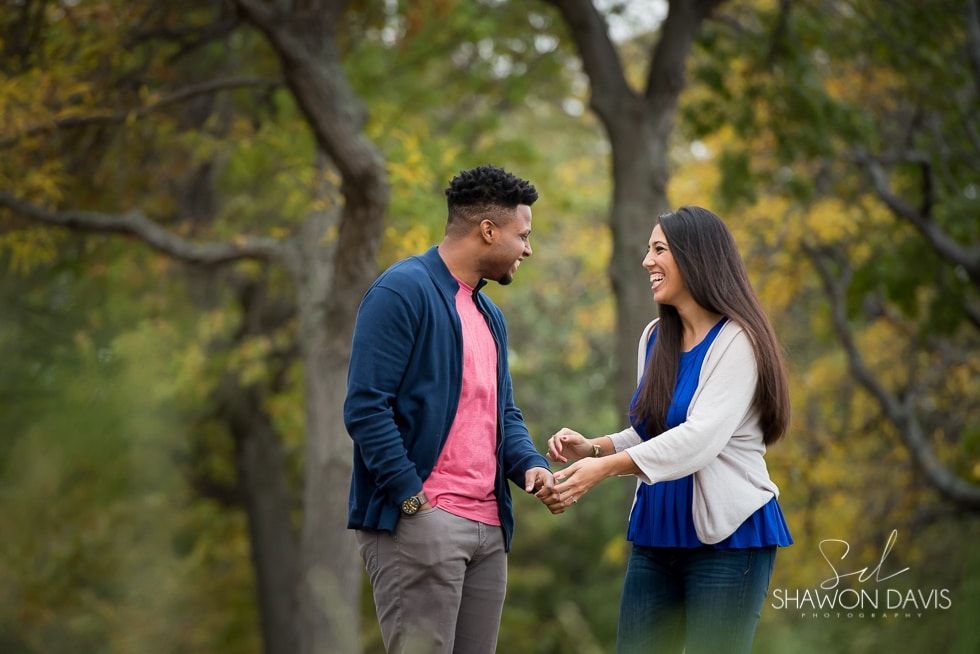 Couple holding hands and smiling in a park with trees and soft greenery.