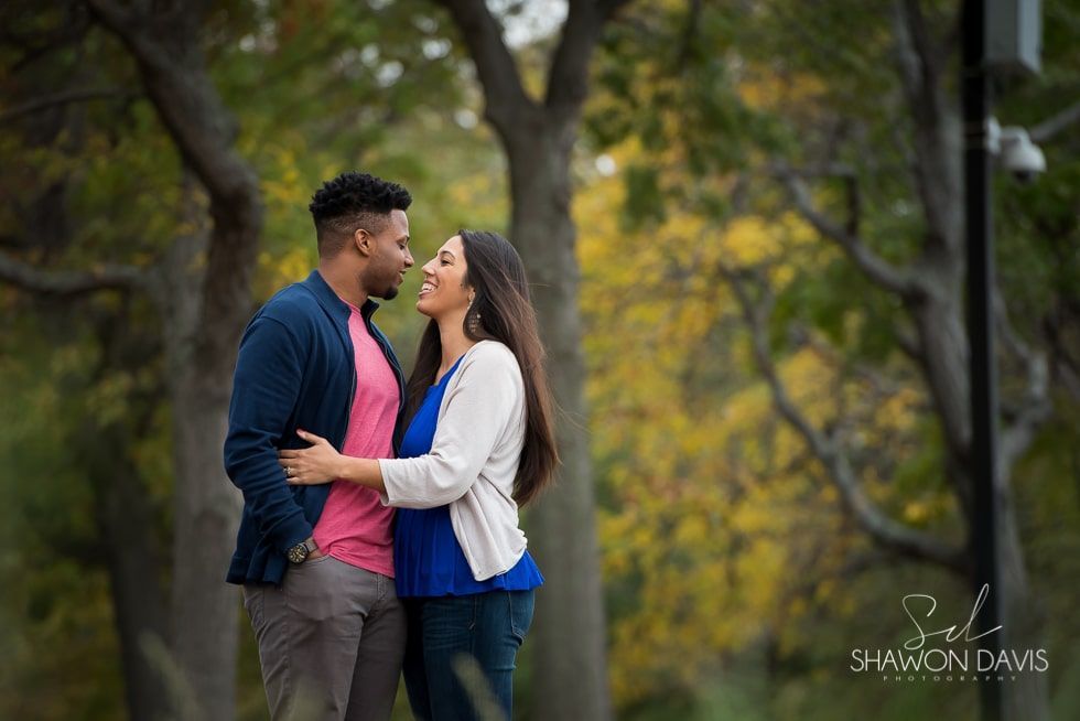 Couple standing close and holding each other in a wooded park, facing one another and smiling