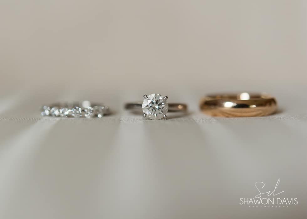 Three rings on a white surface: a silver band, a diamond solitaire, and a gold band.
