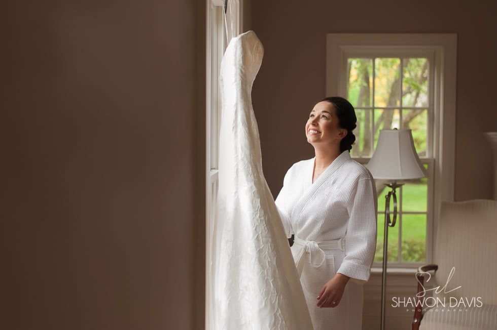 Woman in a white robe smiling beside a wedding dress in a softly lit room
